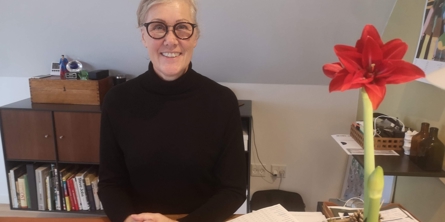 Portræt af ejer af Rehaps Heidi Siegvart, der sidder bag et skrivebord med hænderne foran sig hvilende på bordet. På bordet står en opslået bærbar computer og en potteplante med rød blomst