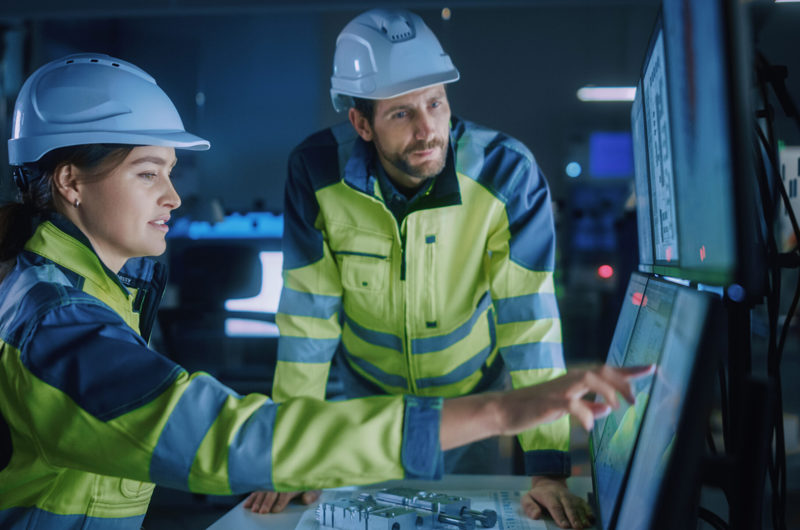 Mand og kvinde iført hvide sikkerhedshjelme og gule refleksjakker kigger sammen på en computerskærm, hvor kvinden peger på noget