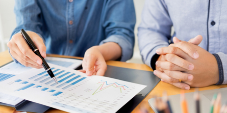 Nærbillede af to personers torsoer. Personerne, der er klædt i blå skjorter, sidder ved et bord, og den ene peger med en kuglepen på nogle grafer på et stykke papir, hun holder. Den anden person har foldede hænder.