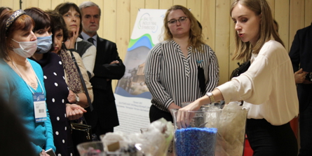 En flok mennesker kigger på en kvinde, der taler, mens hun har hænderne nede i en glasbowle med blåt granulat