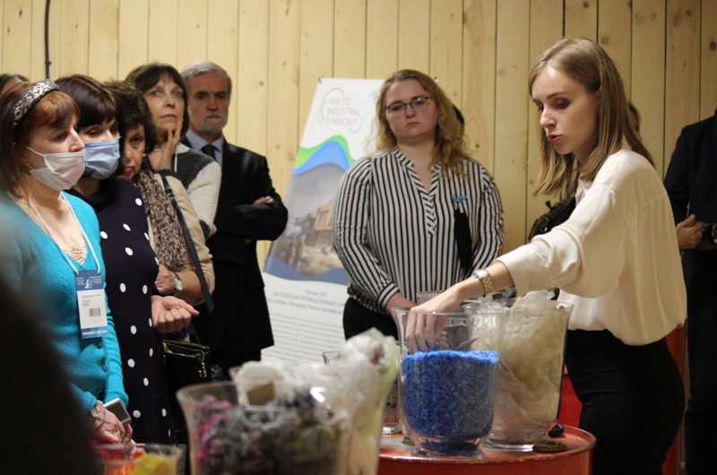 En flok mennesker kigger på en kvinde, der taler, mens hun har hænderne nede i en glasbowle med blåt granulat