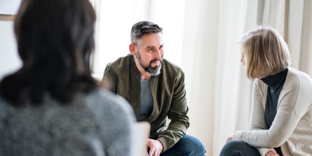 To personer sidder i koncentreret samtale, mens en tredje med ryggen til beskueren ser ud til at lytte med