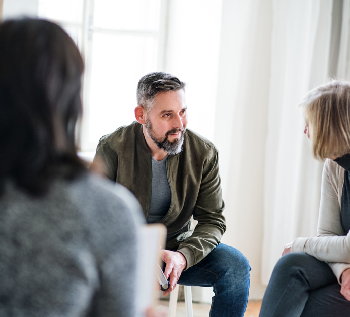 To personer sidder i koncentreret samtale, mens en tredje med ryggen til beskueren ser ud til at lytte med