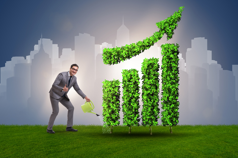 Mand i grå habit vander med vandkande på en opadgående kurve lavet af grønt græs, mens han vender tommelfingeren op til fotografen