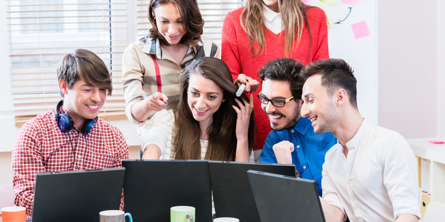 Seks glade unge i muntre farver med meget rødt står og sidder bag et bord, og kigger på en computerskærm foran dem