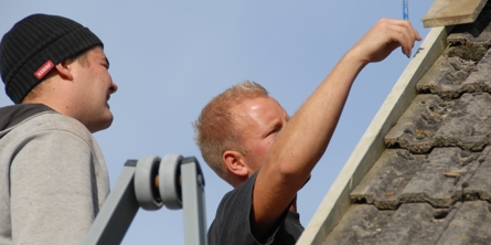 Nærbillede af to mænd, der står på stiger ved et hustag. den ene er i gang med at måle noget op.