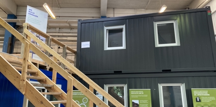 To sorte containere med vinduer i siden står stablet ovenpå hinanden. I forgrunden fører en trappe i lyst træ op til et plateau foran den øverste container