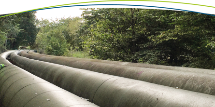 En række store rør løber gennem et skovlandskab. Ved siden af dem med ryggen til beskueren går to mænd iklædt gule refleksjakker